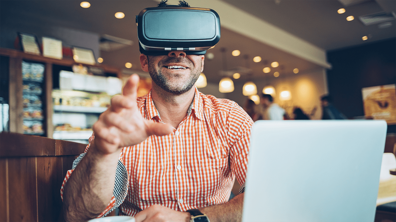 Smiling man in cafe with laptop and VR headset reaches for an unseen object.