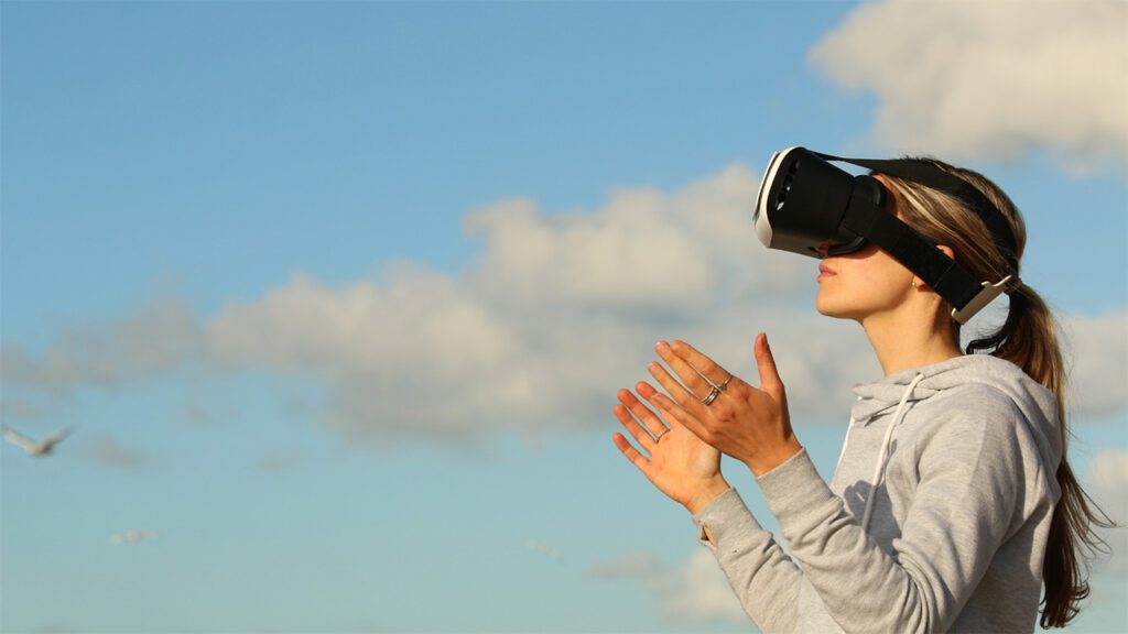 A woman wears a VR headset. In the background a blue sky and soft clouds.