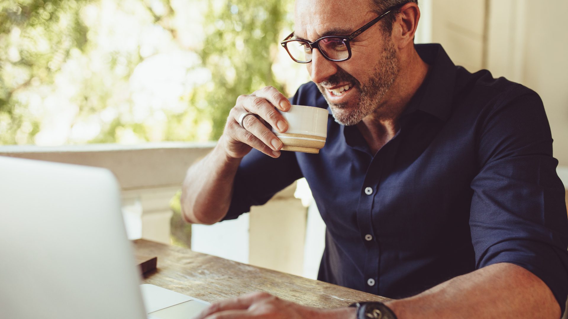 Middle-aged man with glasses, raises a small coffee cup to his mouth while looking at a laptop and smiling.