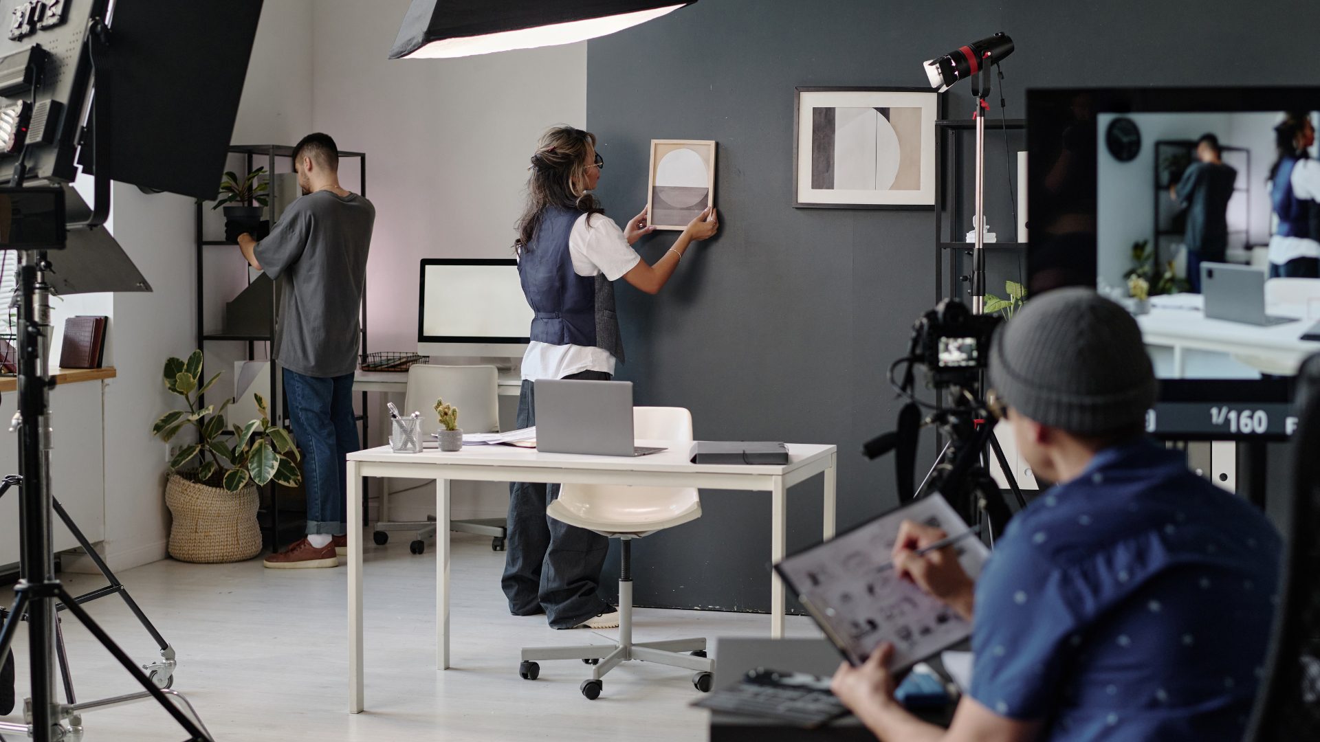 On set. A production team dresses the set with an art director reviewing the storyboard on a clipboard. In the foreground we see tripods with cameras and lighting equipment.