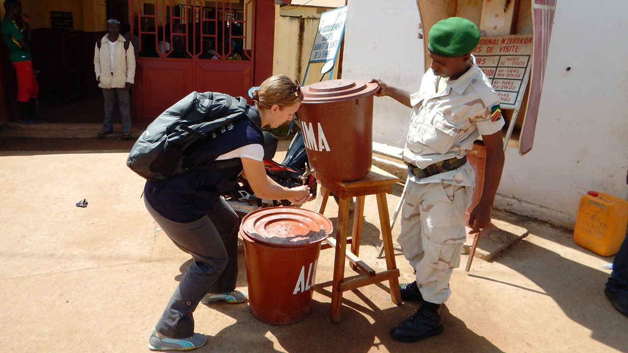 A woman washes her hands at a makeshift washing station while uniformed soldier looks on.